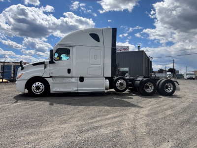 Truck side view showing a white Freightliner CA-126 tractor with sleeper cab and multiple rear axles on a gravel lot under blue sky.