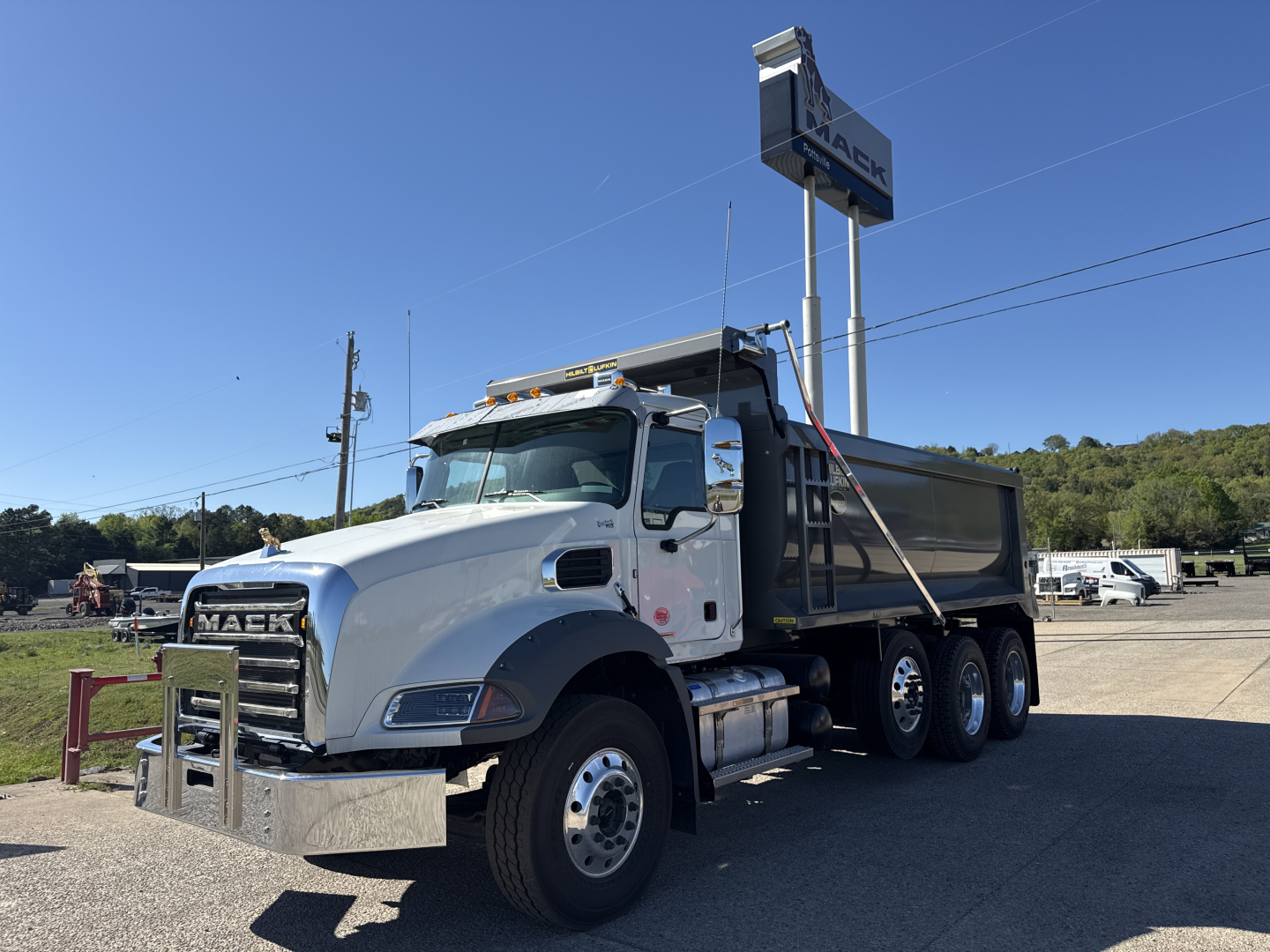 Dump truck with Mack grille showing three-axle configuration and dump bed, chrome bumper, side mirrors, and steel chassis; visible signage in lot.