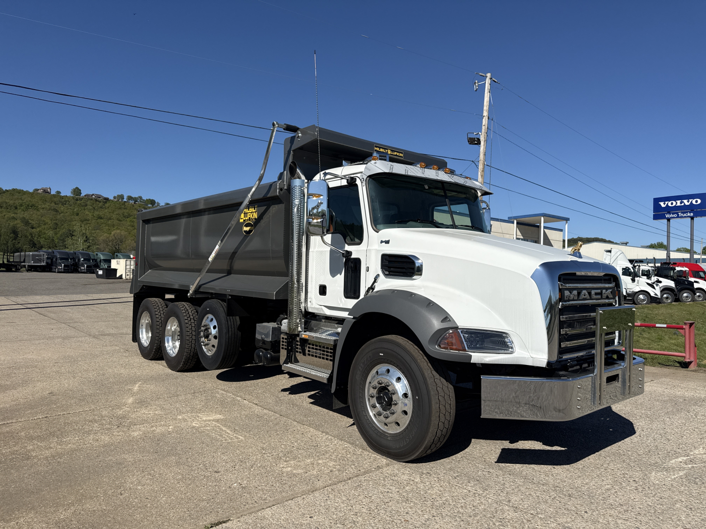 Dump truck in white and black with a large grille, six wheels, and a dump bed. Visible text includes Mack grille and truck body details, showing a heavy-duty dump truck from the front-left angle.