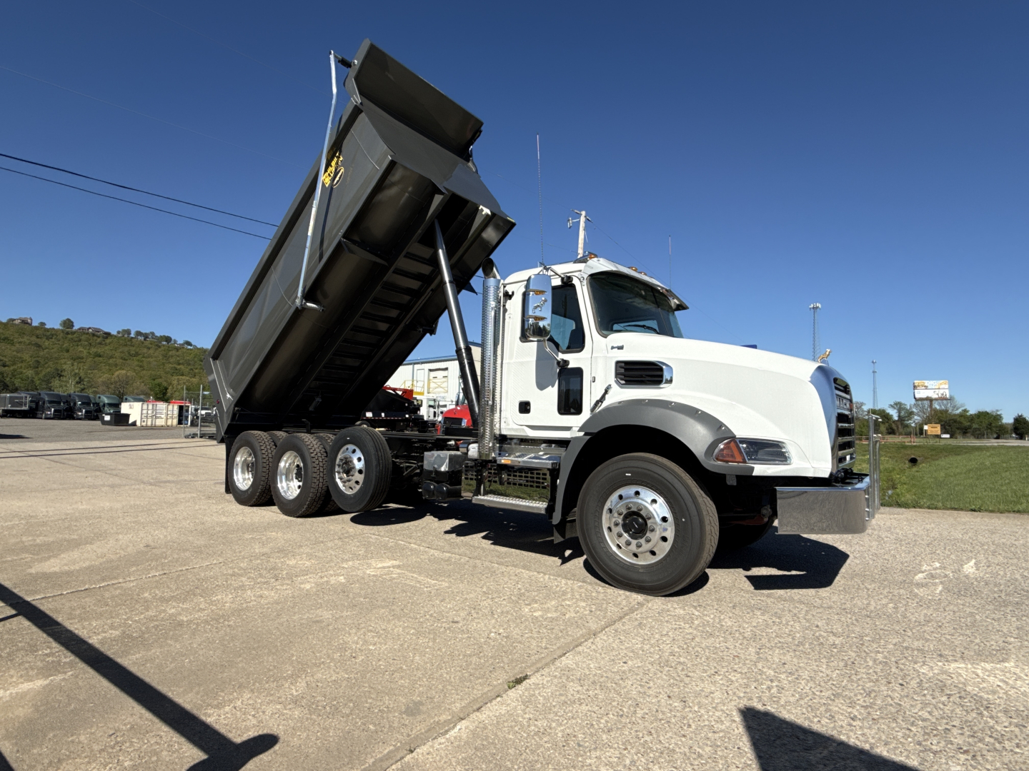 Dump truck with raised dump bed on a white Mack GR84B with three-axle configuration, front cab chrome bumper, side view showing vehicle details.