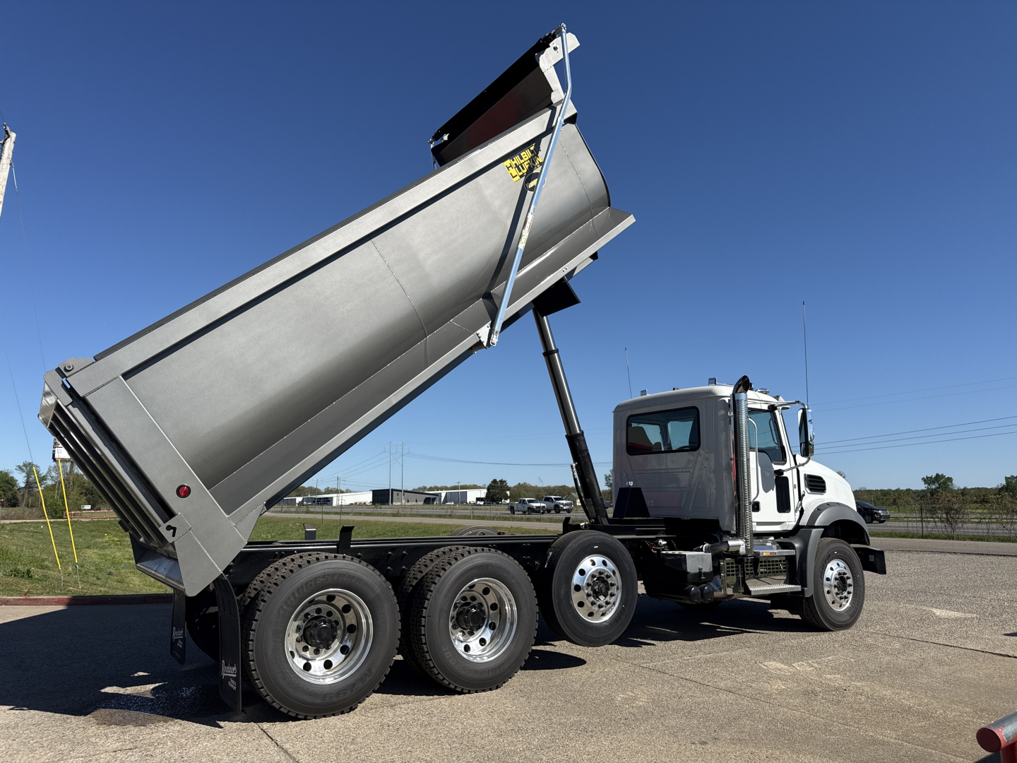 Dump truck with raised dump bed on Mack GR84B showing triple rear axles and hydraulic lift under a clear blue sky.