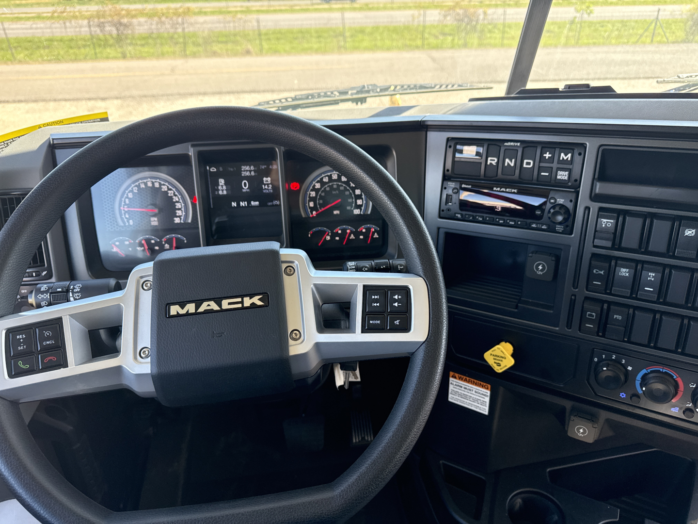 Mack GR84B truck interior showing steering wheel with Mack logo, dashboard with gauges, digital display, and control panel including radio and HVAC knobs.
