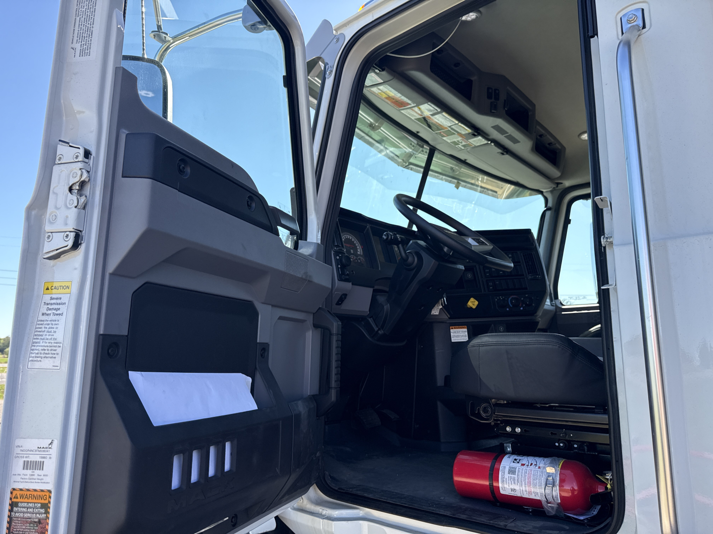 Truck cabin of 2026 Mack GR84B showing open door, steering wheel, dash, and a red fire extinguisher on the floor.