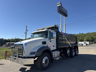 Dump truck with Mack grille showing three-axle configuration and dump bed, chrome bumper, side mirrors, and steel chassis; visible signage in lot.