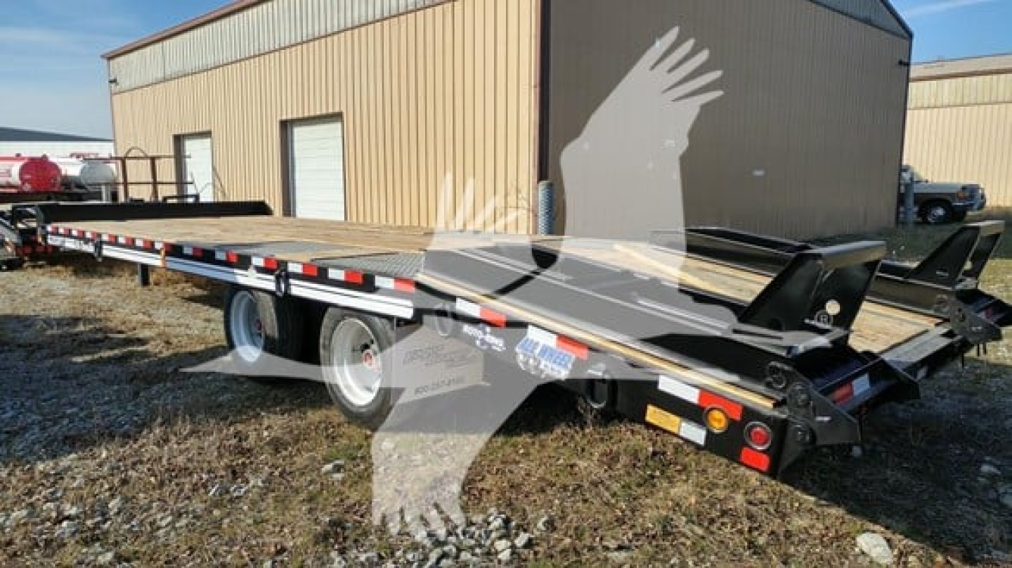 Flatbed trailer in yard showing long deck, rear ramps, and dual axles with white wheels.