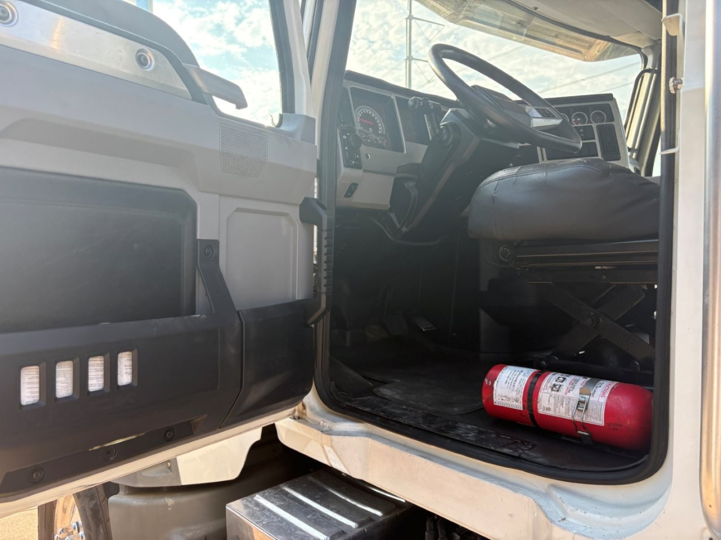 Cab interior of a 2021 Mack GR64F dump truck showing the driver seat, steering wheel, dashboard gauges, and a red fire extinguisher on the floor near the door.