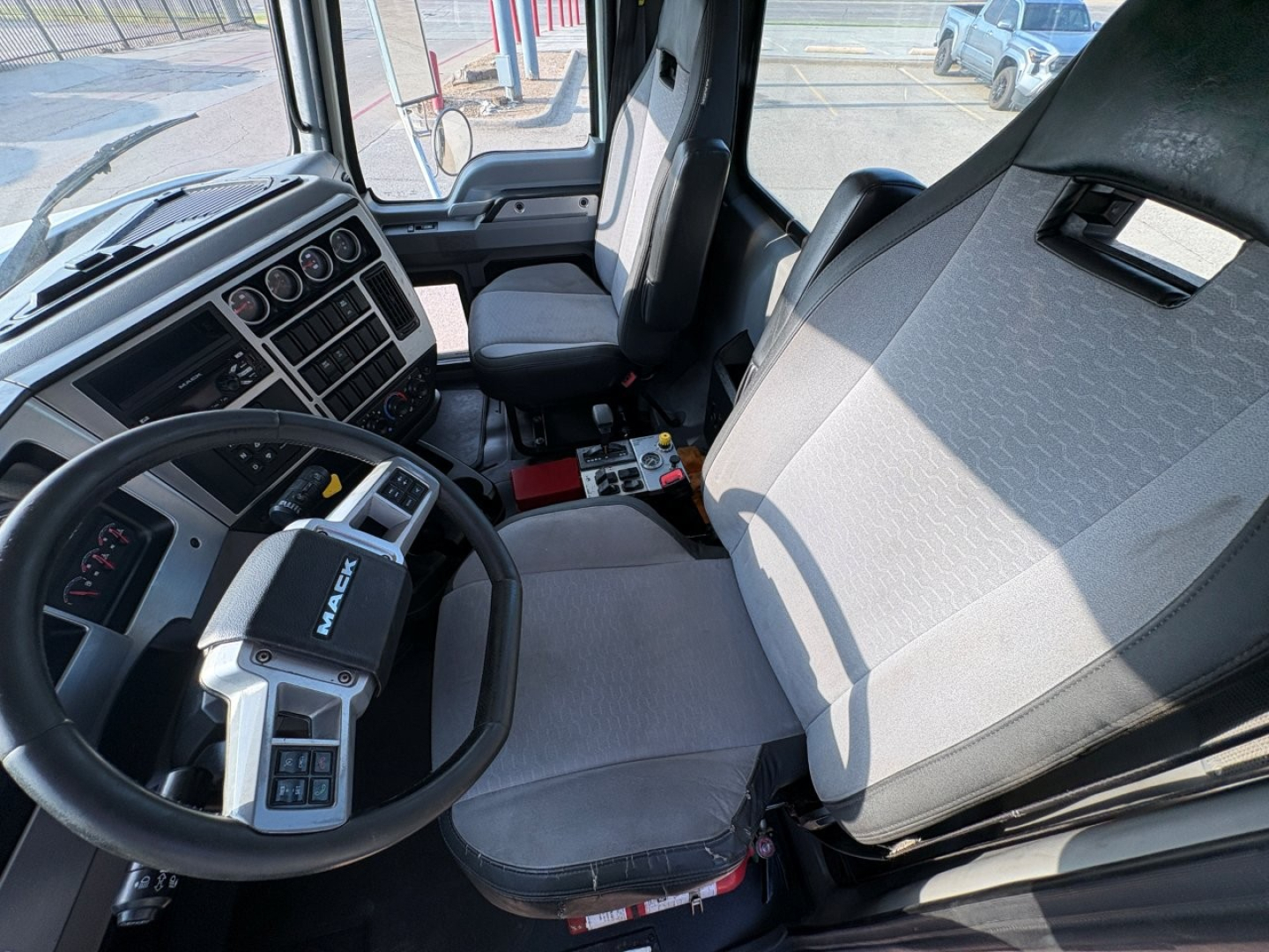 Truck cab interior of a 2021 Mack GR64F showing steering wheel, dashboard gauges, driver seat, and passenger seat with center console“, showing utility controls and standard cab layout.
