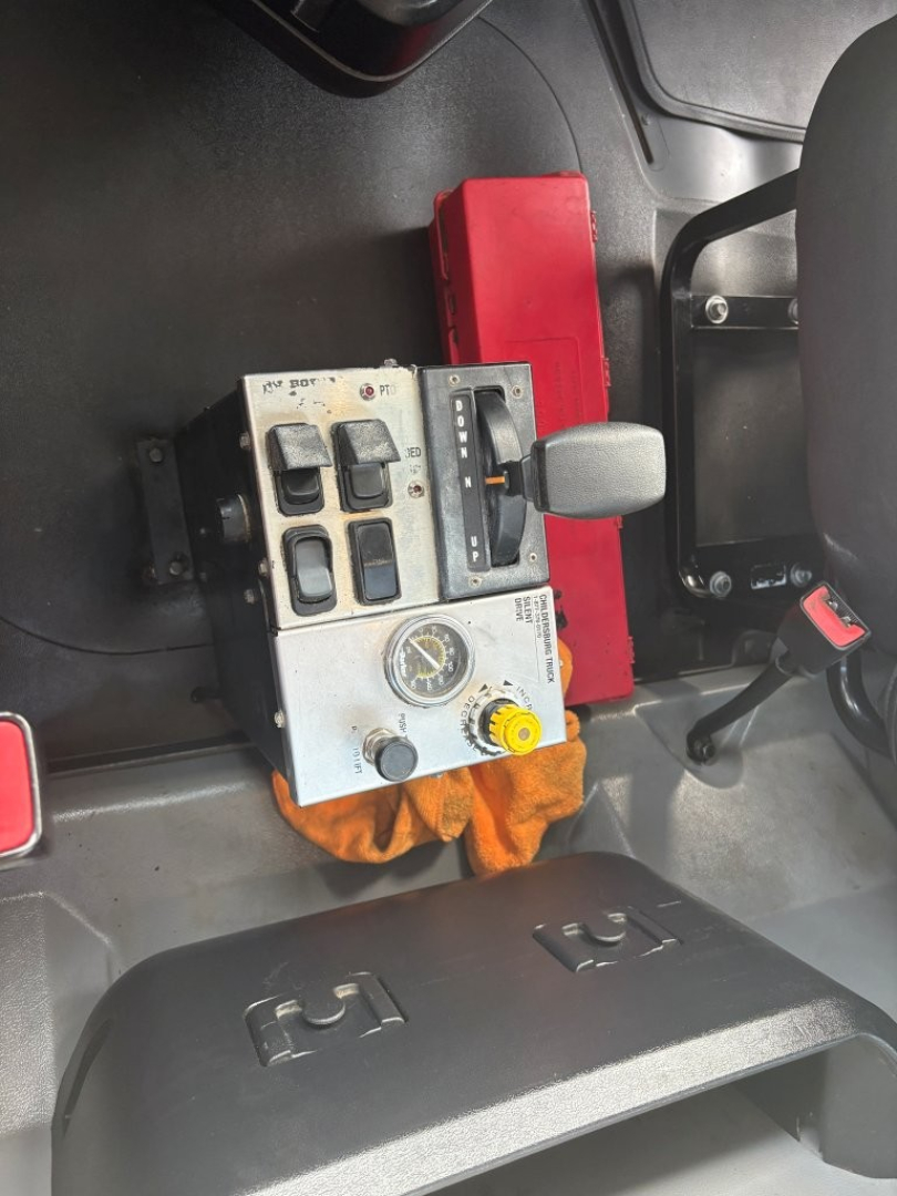 Truck cab interior with control panel including multiple switches, gauges, and a yellow knob; gear lever nearby; red box and orange cloth visible.