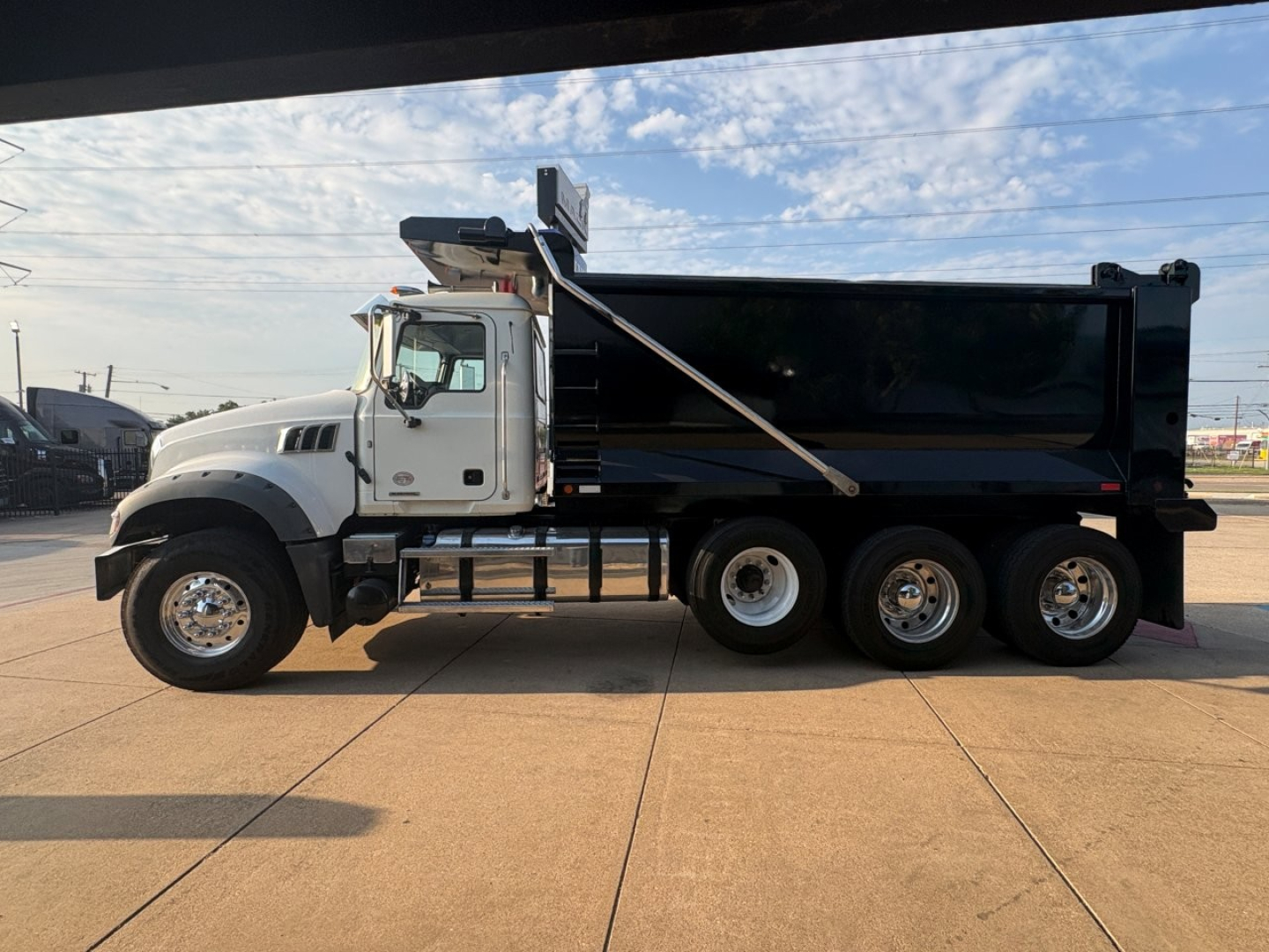 2021 Mack GR64F dump truck side view showing large black dump bed, white cab, three rear axles, chrome wheels, and hydraulic mechanism.