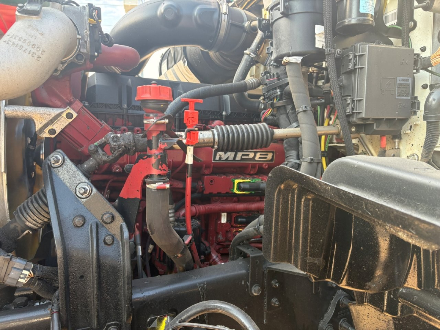 Mack GR64F engine bay showing red MPI 8? engine with black intake, hoses, and wiring harness; visible dipstick and surrounding components in service bay.