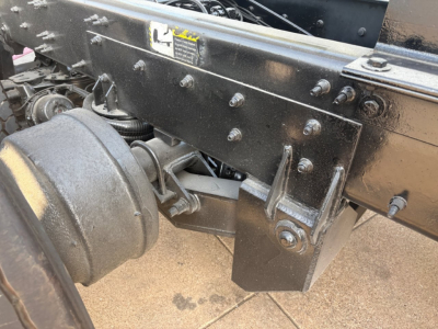 Mack GR64F chassis view showing suspension hanger and frame with bolts and a leaf spring mount area. Visible components include a large drive axle housing and frame crossmember with bolts.