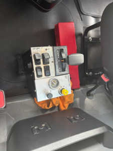Truck cab interior with control panel including multiple switches, gauges, and a yellow knob; gear lever nearby; red box and orange cloth visible.