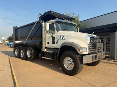 Mack GR64F dump truck in-service showing front grille, cab, and side of dump bed with elevated hydraulic hoist and chain,”