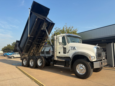 Truck: 2021 Mack GR64F dump truck with raised dump bed, white cab, black dump body, three rear axles, visible VIN plate area. Side view showing wheel setup and hydraulic lift.