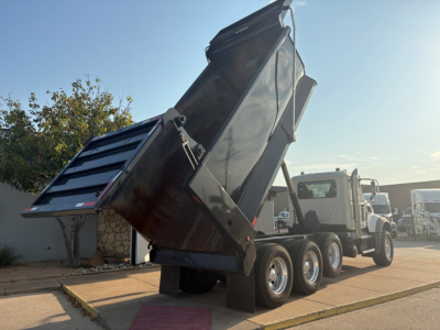Dump truck in-service Mack GR64F with raised dump bed and triple rear axles, side view showing hydraulic mechanism and cargo bed.