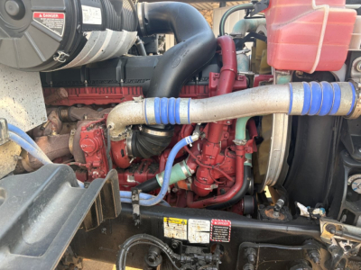 Mack GR64F engine bay showing red turbo diesel engine with blue silicone hoses, radiator cap, coolant reservoir, and label stickers visible.