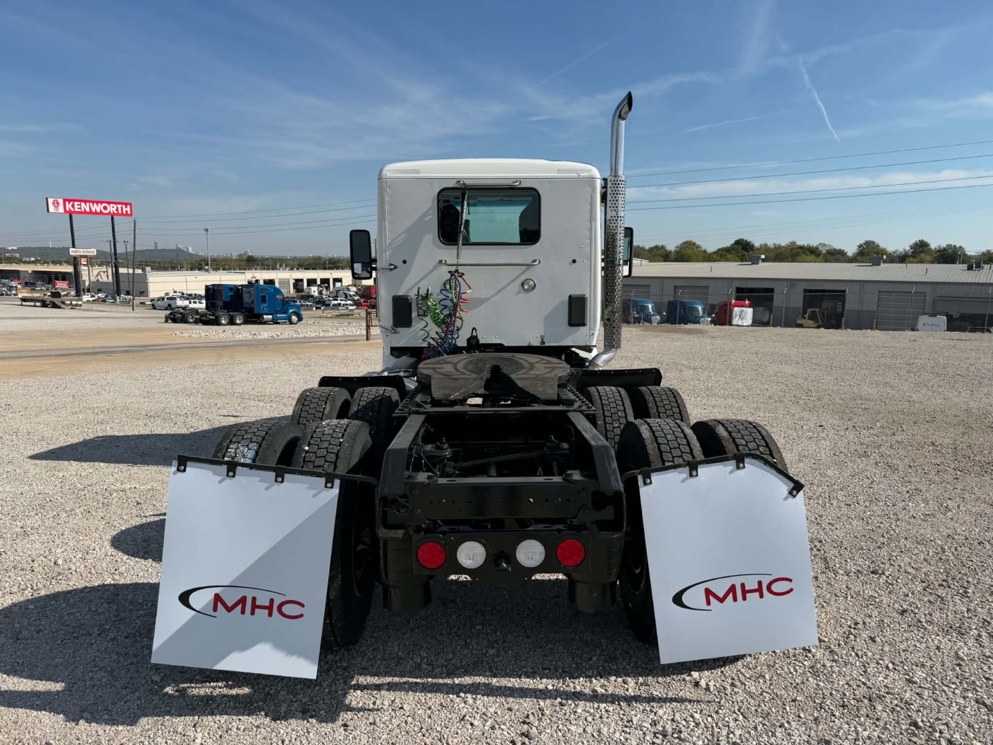 Kenworth T880 rear view showing mud flaps with MHC logos, fifth wheel, and dual rear axles in a gravel lot.