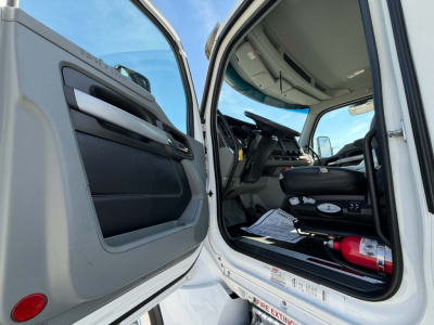 Kenworth T880 cab interior with open driver door showing steering wheel, dashboard, and seat; a fire extinguisher secured on the floor near the door.