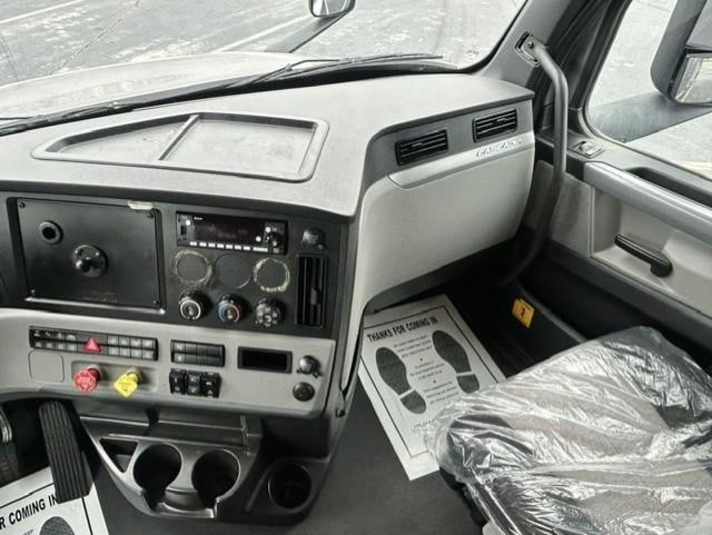 Truck cab dashboard of a 2022 Freightliner Cascadia showing instrument cluster, center console, and driver seat; dashboard panels with controls and vents; plastic protective cover on seat.
