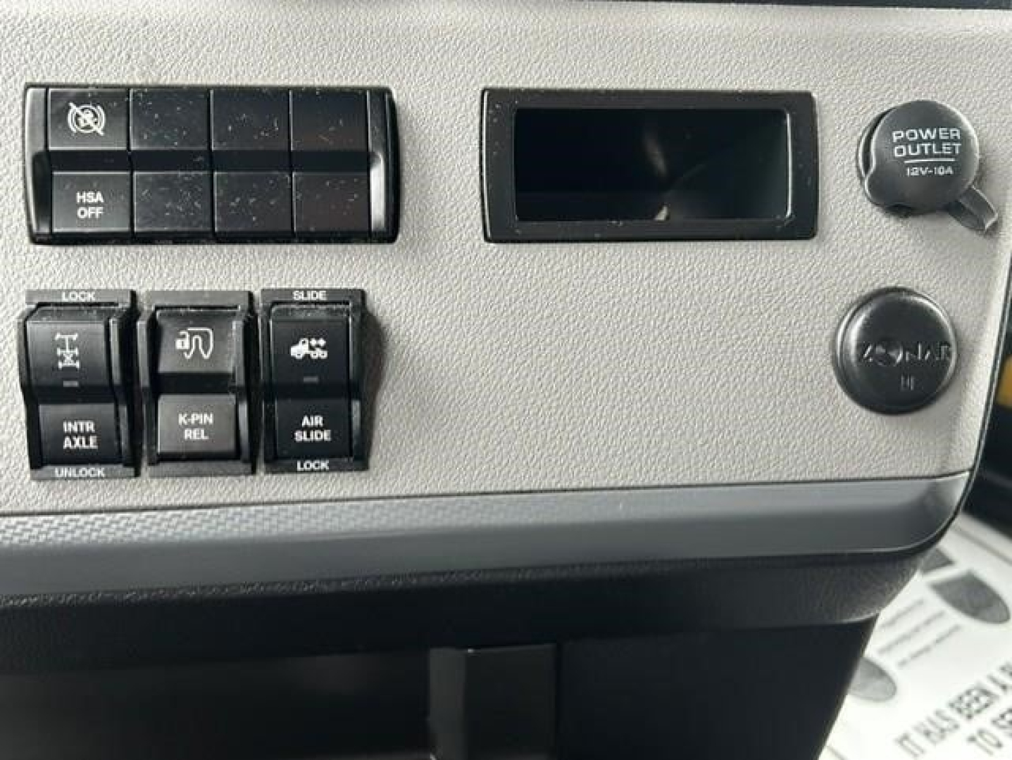 Dashboard controls of a Freightliner Cascadia showing auxiliary switches, power outlet knob, and blank display slot.