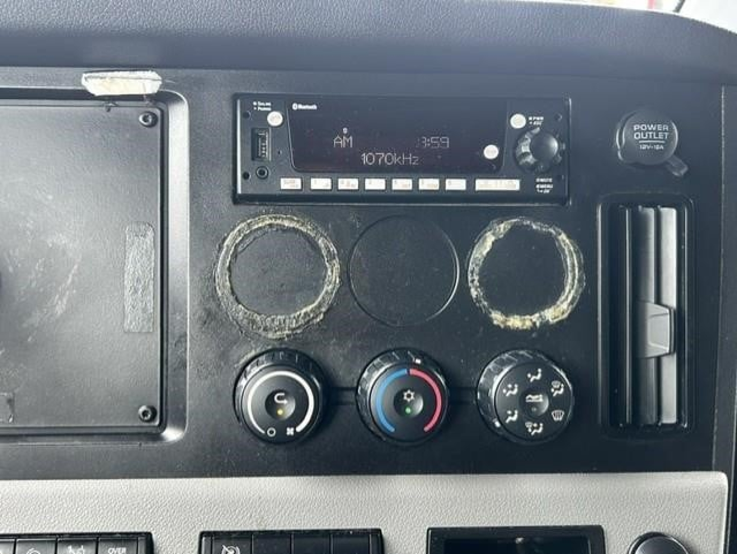 Dashboard panel of a Freightliner Cascadia showing radio unit, three circular gauges, HVAC controls, and power outlet.