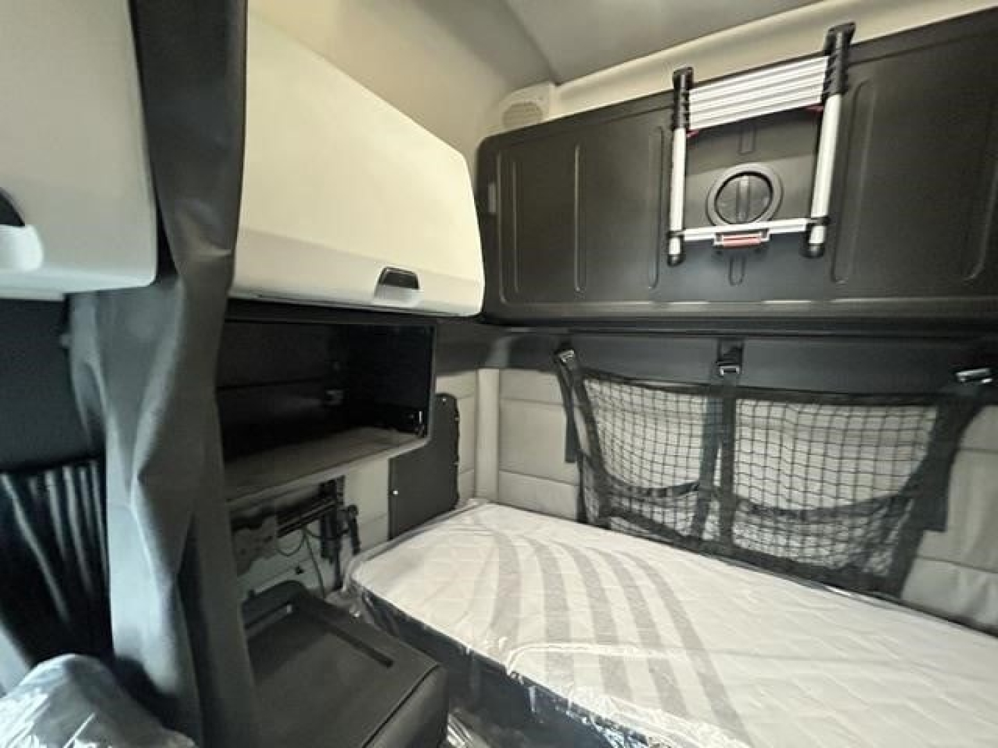Truck sleeper interior with bunk bed area in a Freightliner Cascadia cab; storage shelves, netting on wall, and a mounted device panel visible.