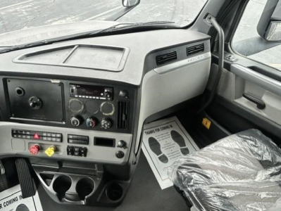 Truck cab dashboard of a 2022 Freightliner Cascadia showing instrument cluster, center console, and driver seat; dashboard panels with controls and vents; plastic protective cover on seat.