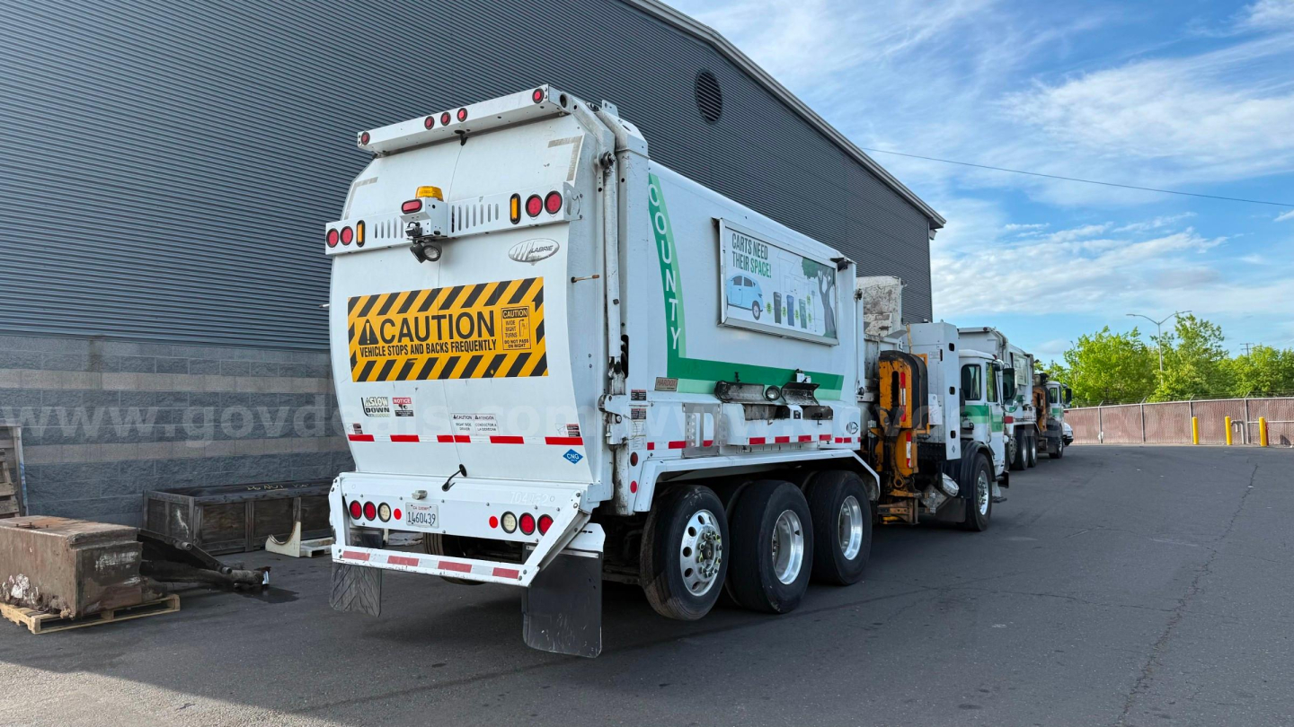 Garbage truck in service position showing rear view of waste collection unit with caution sign and multiple axles.