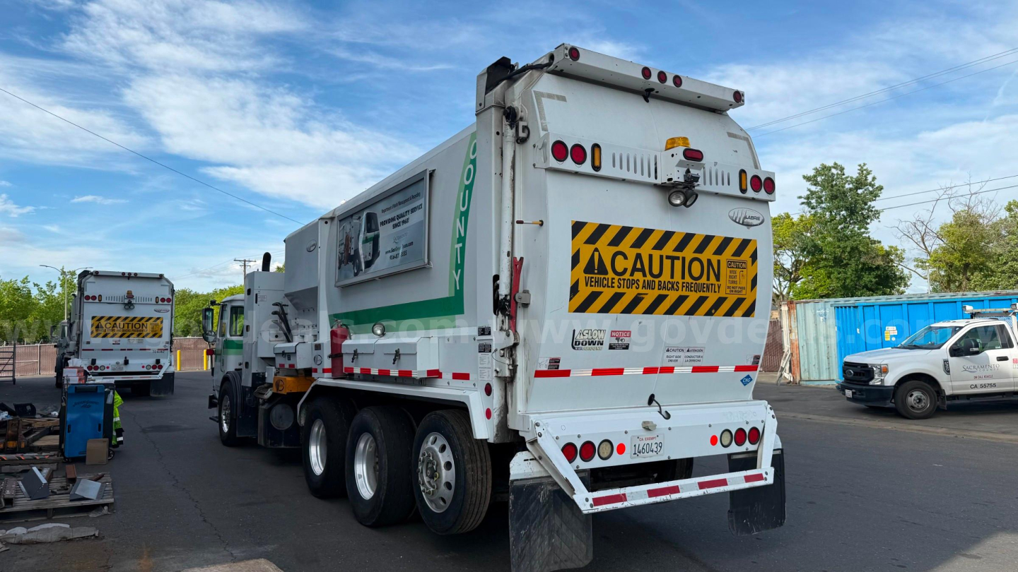 Rear view of a white Autocar garbage truck with CAUTION sign, multiple rear lights, and a green panel on the side showing 'GON' or similar. The truck has triple axles and a refuse compactor body with safety decals.