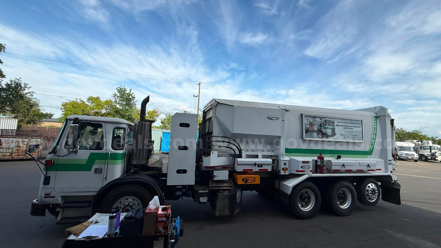 Autocar garbage truck side view showing a green-and-white cab and a large gray rear body with wheel group and hydraulic components.