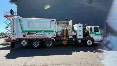 Autocar ACX XPeditor side view garbage truck with green and white livery, tank bed, and recycling chute. Visible text on side reads: 'COUNTY' vertical on rear, and a large silver access panel with small text. Cab and truck show typical heav