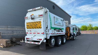 Garbage truck in service position showing rear view of waste collection unit with caution sign and multiple axles.