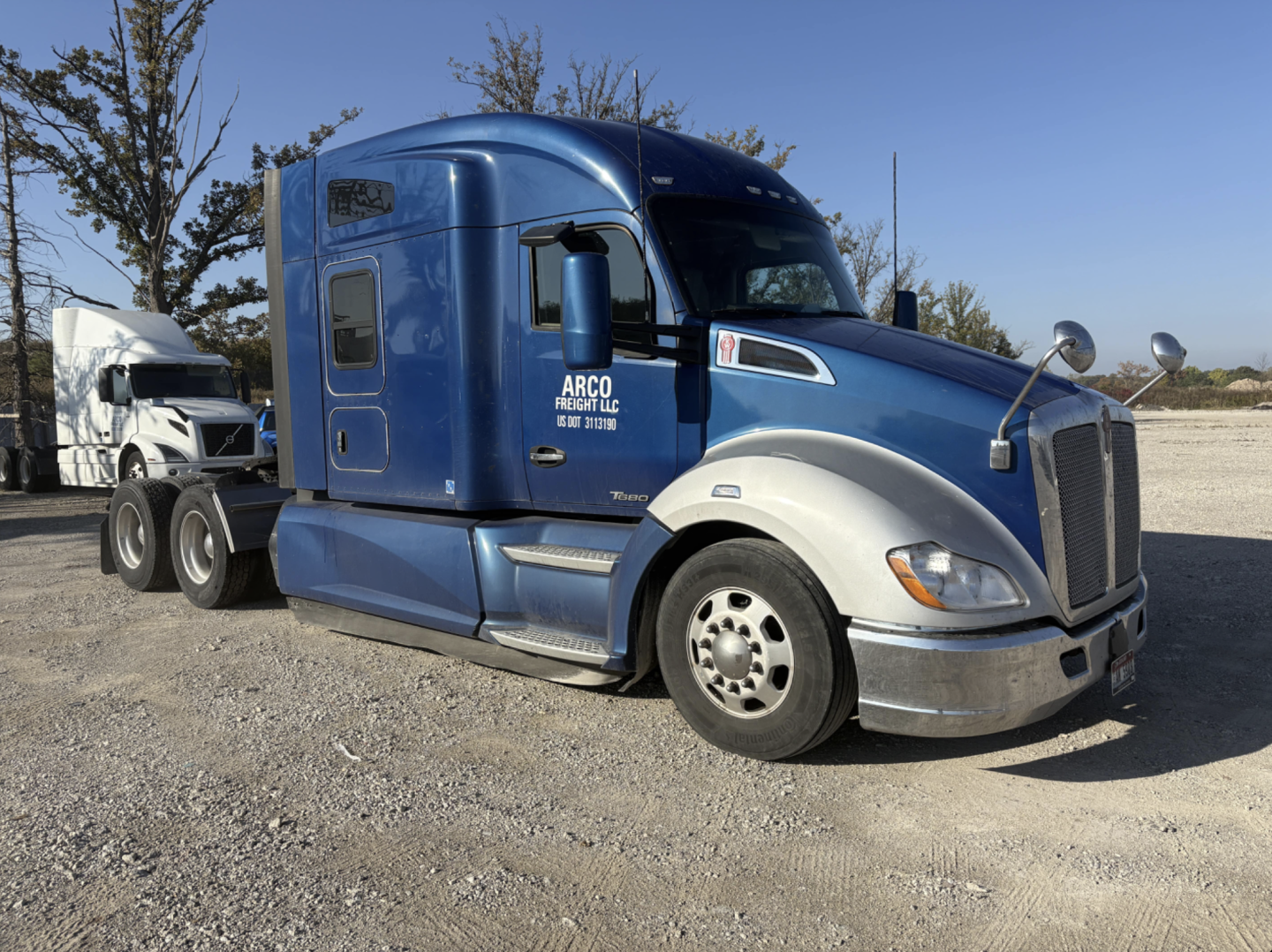 Kenworth T680 sleeper cab tractor in blue and silver, side view with front grille, mirrors, and ARCO Freight LLC decal; highway truck with a secondary trailer in background.