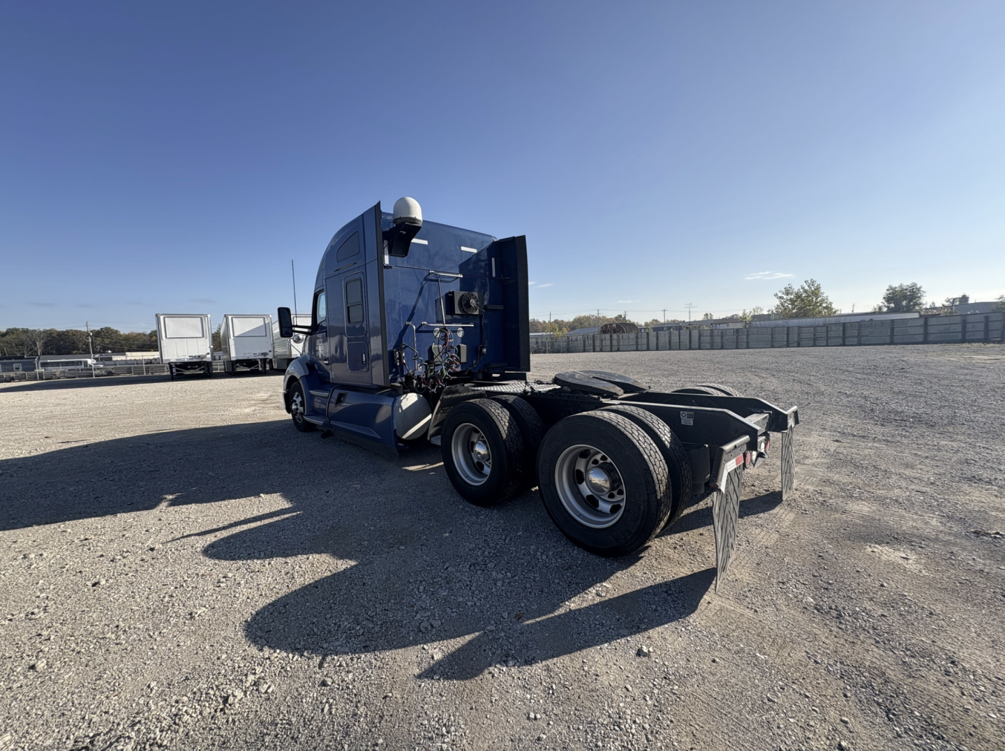 Kenworth T680 semi truck tractor chassis view from rear left, with third and fourth wheels visible, blue cab, gravel lot, clear sky.