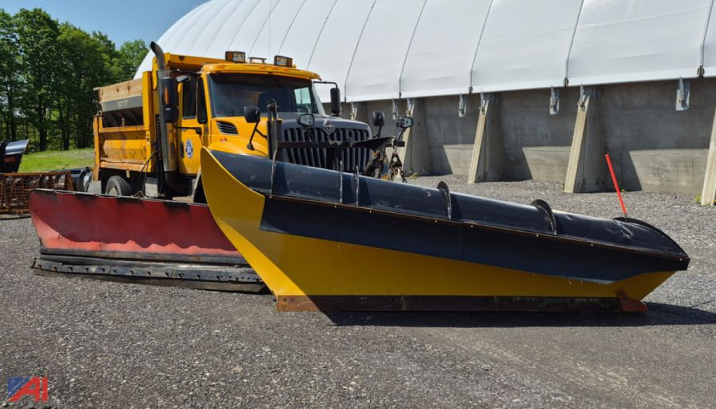 Heavy-duty orange International WorkStar 7600 dump truck with a large yellow and black snow plow wing attachment in front, parked beside a white dome building.
