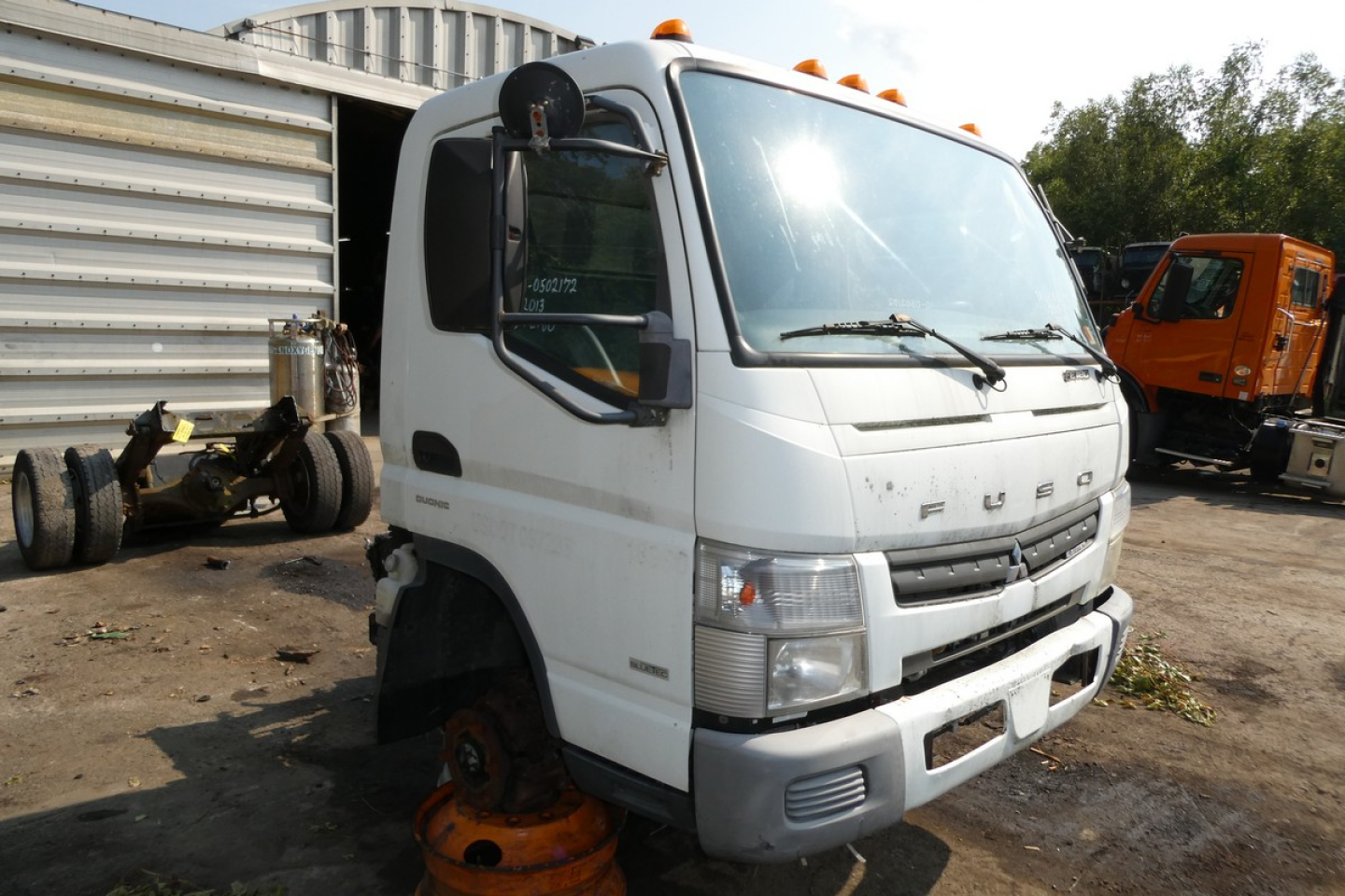 Mitsubishi Fuso FE180 cab and chassis truck in a yard, front view with missing front wheel and orange jack stands nearby, visible cab and windshield.