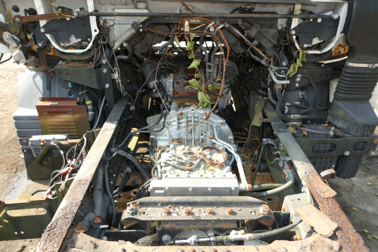 Front view of a Mitsubishi Fuso FE180 chassis with exposed drivetrain; transmission mounted between frame rails, wiring harness, and rusted rails visible.