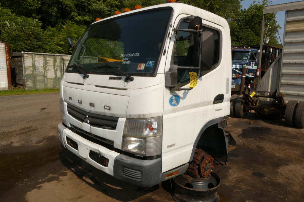 Mitsubishi Fuso FE180 cab and chassis truck front view showing removed front wheel and blank wheel hub, white cab with black grille and headlights.