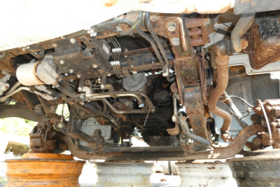 Underbody view of a 2013 Mitsubishi Fuso FE180 chassis showing tangled hydraulic lines and rusted metal frame components.