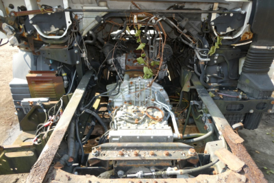 Front view of a Mitsubishi Fuso FE180 chassis with exposed drivetrain; transmission mounted between frame rails, wiring harness, and rusted rails visible.