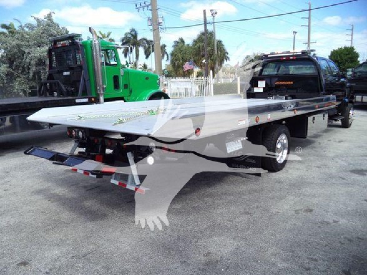 Flatbed rollback tow truck in driveway with green heavy-duty tow unit and a black pickup on a dual-wheel trailer bed. Visible VIN tag and registration stickers.
