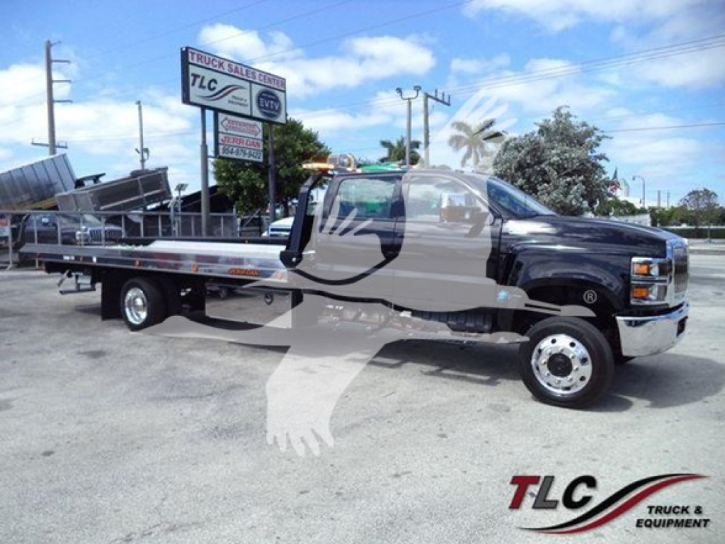 Flatbed tow truck with cab showing International branding and long metal flatbed. Visible sign in background with TLC Truck Sales. Vehicle shows VIN sticker and stock number J19807 area not legible.