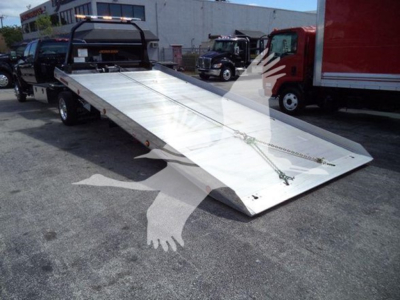 Truck rollback tow bed with extended flatbed showing metal surface and ramp in front; chain tied to side and a red box truck in background.