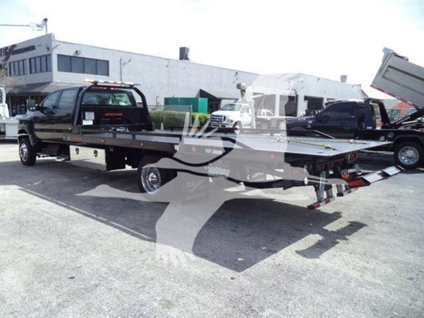 Flatbed rollback tow truck with a black cab and a long flatbed trailer, chrome wheels, hydraulic bed tilt, and adjacent vehicles in a dealership yard.