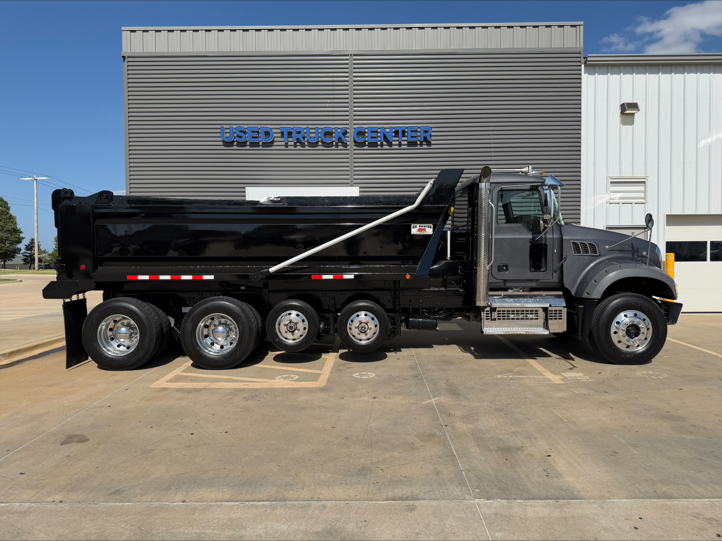 Dump truck side view of a 2023 Mack GR64F showing a black dump bed, multiple axles, metal steps, and cab with gray body; background front of Used Truck Center building; VIN visible on listing.