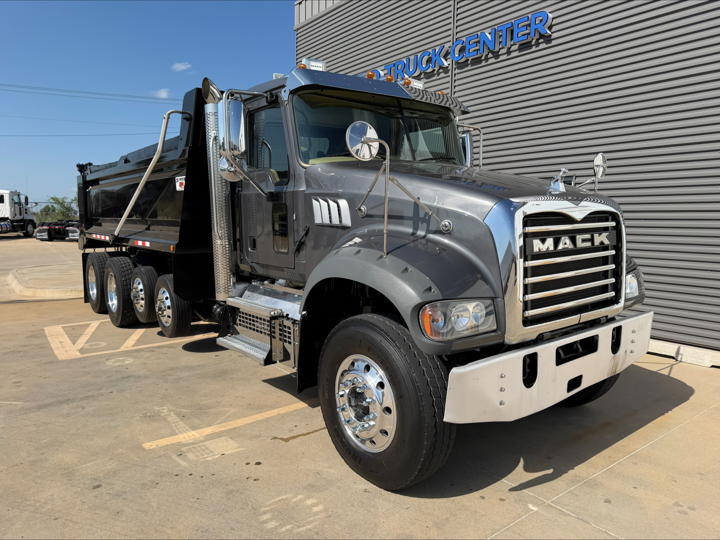 Mack GR64F dump truck on lot, front three-quarter view showing chrome grille with Mack logo, dual headlights, large bumper, black dump bed, and multiple rear axles.