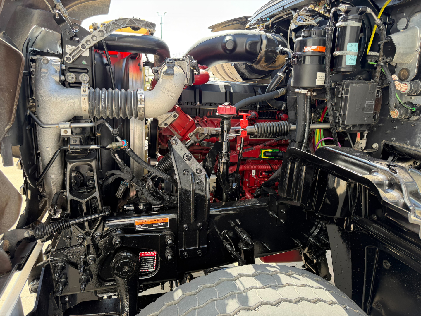 Mack GR64F engine bay showing air intake, hoses, fluid reservoirs, and engine block with red components; visible wiring and surrounding chassis.