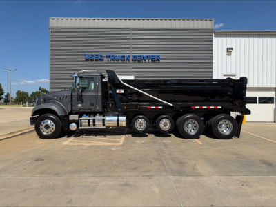 Dump truck (2023 Mack GR64F) side view showing black dump bed, six wheels, and cab with chrome exhausts in front of a building labeled USED TRUCK CENTER.