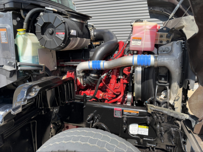 Mack GR64F engine bay showing coolant reservoir, radiator, air filter housing, hoses, and engine block.