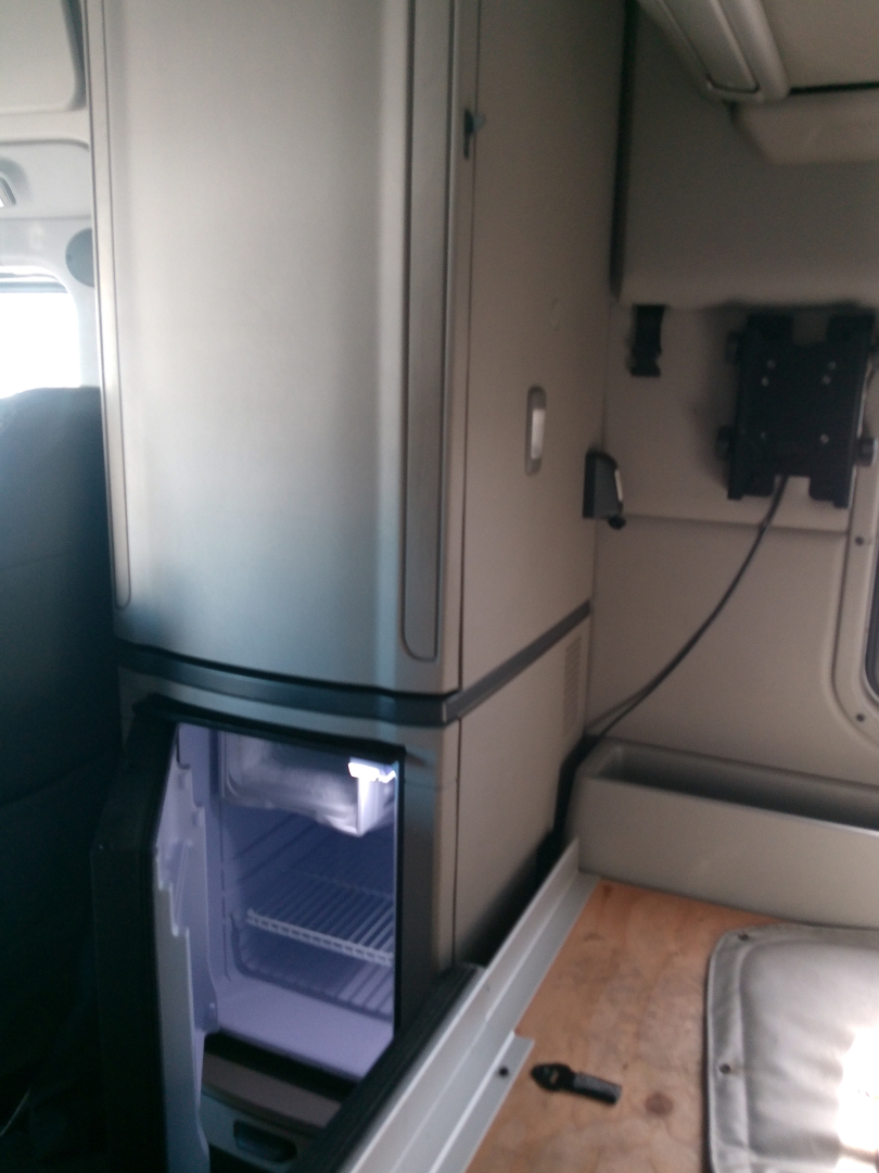 Truck interior showing a vertical refrigerator/freezer unit and a small open compartment; cab area with wiring panel and seating visible.